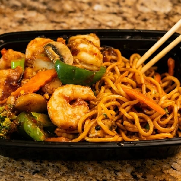 Shrimp and vegetable lo mein with chopsticks in a black takeout container on a granite counter.